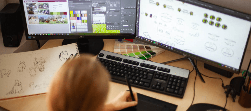 Photo de mon poste de travail, on voit mes écrans d'ordinateur rempli de croquis pour un logo, la planche tendance qui va avec et le bloc de papier avec mes esquisses.